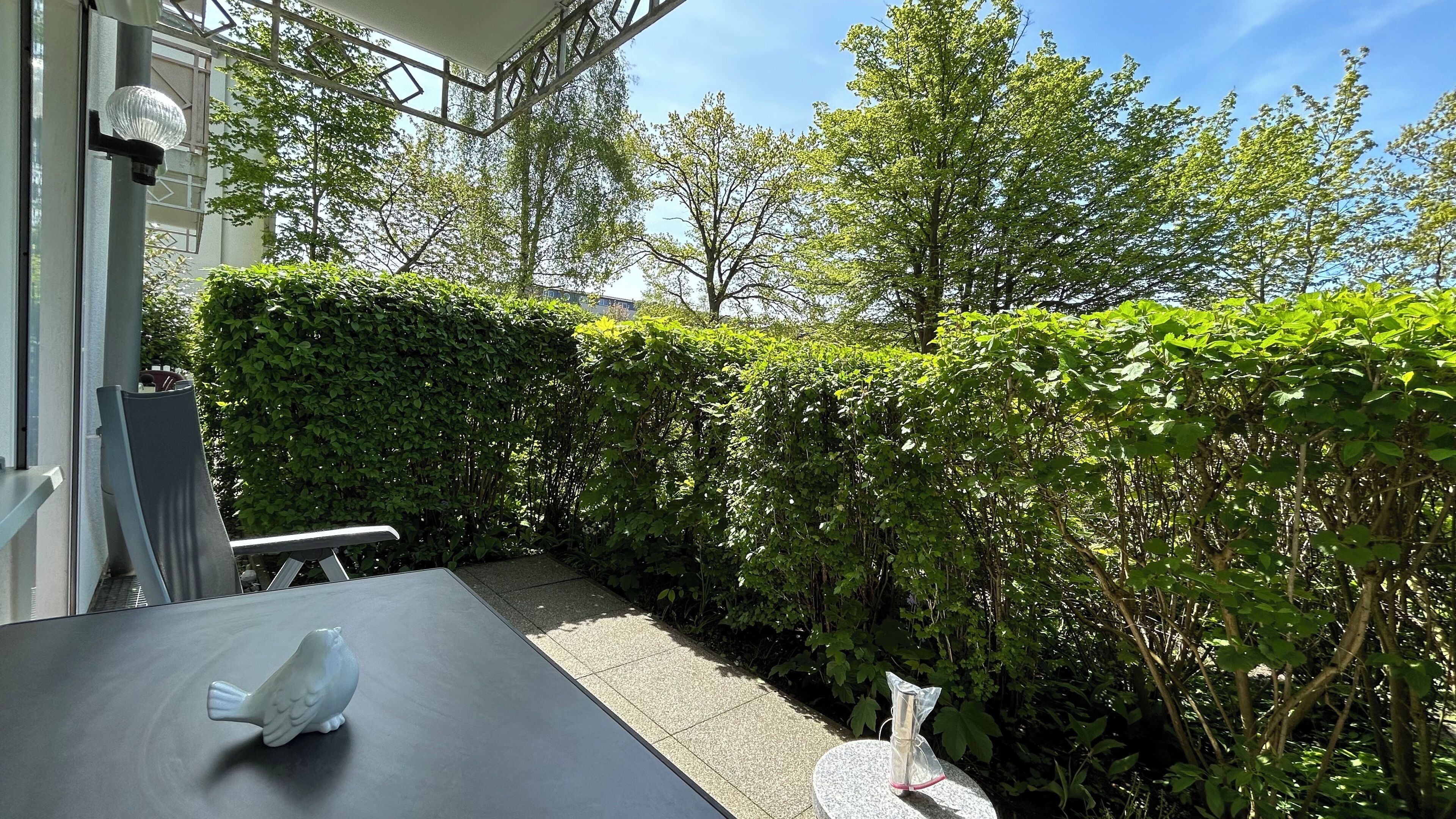 Un'area patio con tavolo e sedie, immersa nel verde e delimitata da un siepe.