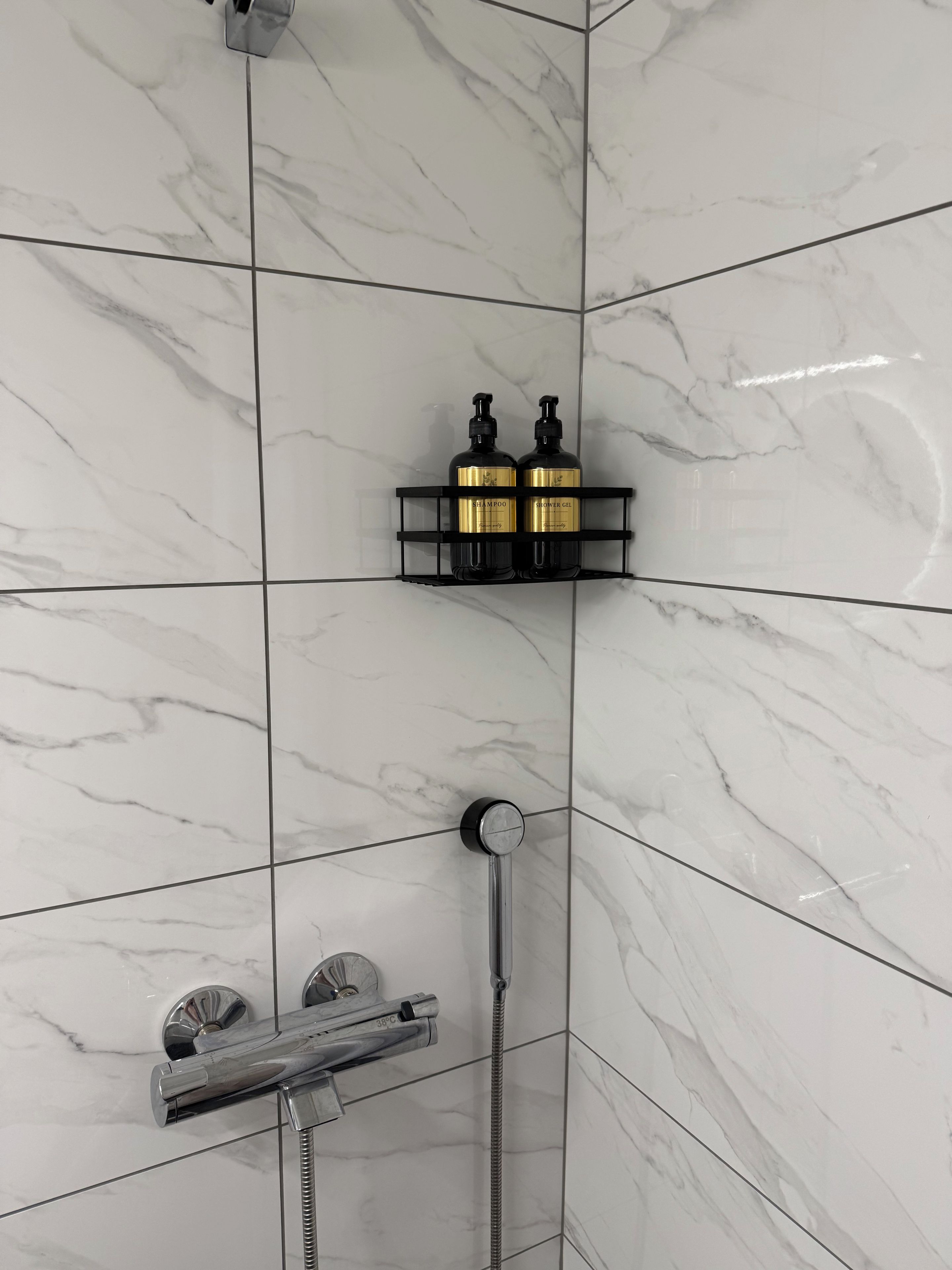 Une salle de bain équipée d’une douche, d’un pommeau de douche à main et d’une étagère noire sur laquelle sont posés deux flacons de shampoing et de gel douche.