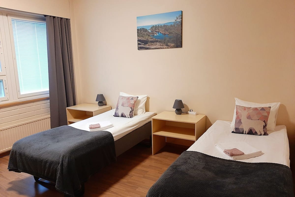 A hotel room with two single beds, a nightstand with a lamp, a small table, a window with blinds, and a painting on the wall.