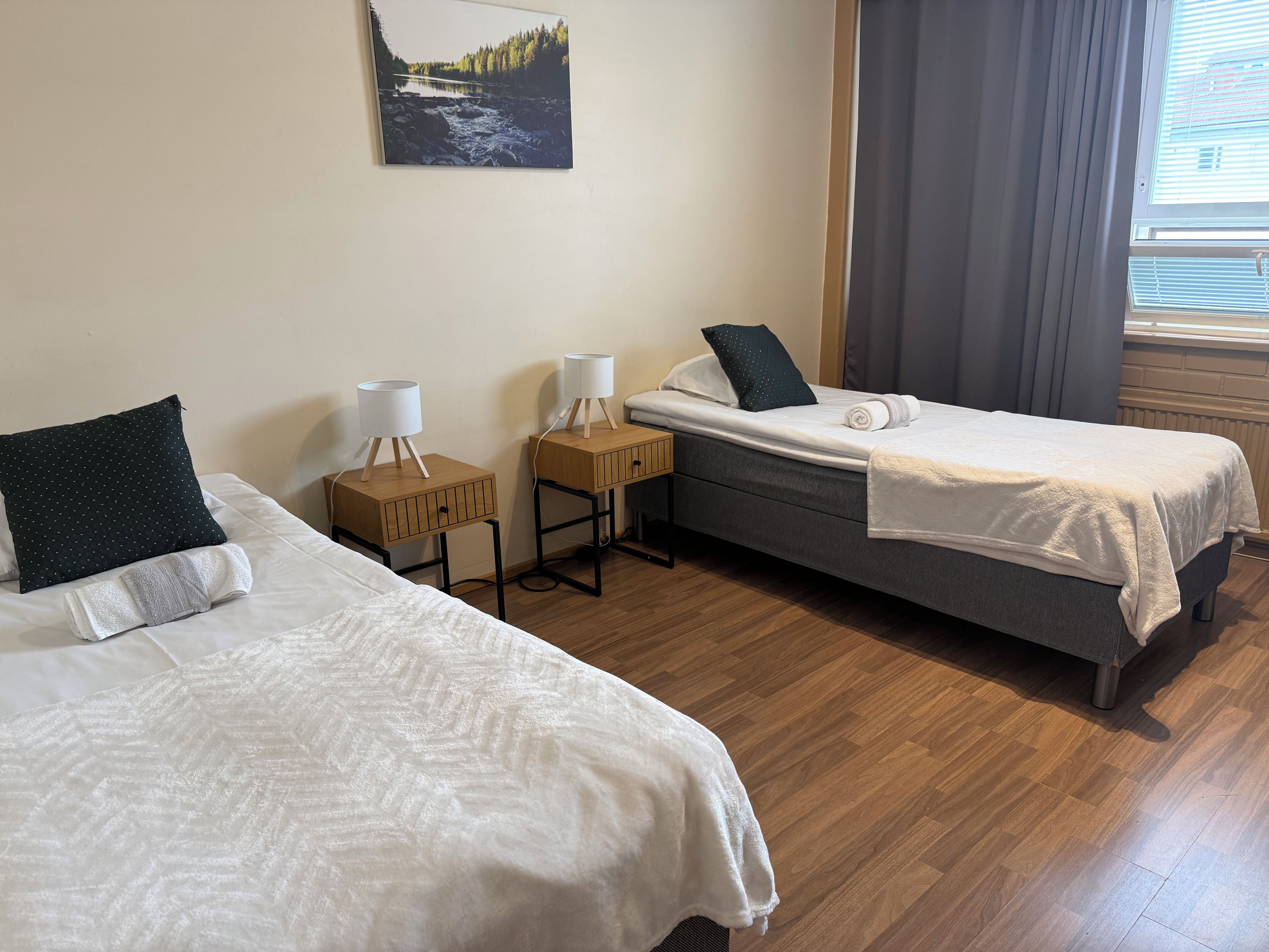 A hotel room with two beds, a nightstand with a lamp, a painting on the wall, and a window with curtains.