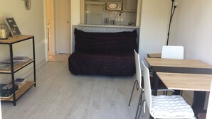 Kompakti olohuone, jossa on tummanvärinen sohva, pieni ruokapöytä tuoleineen, kirjahylly ja taustalla pieni keittiö.
