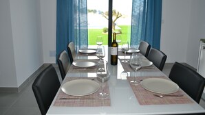 Mesa de comedor lista para seis personas, con platos, copas y una botella de vino, ubicada frente a una ventana con cortinas azules.