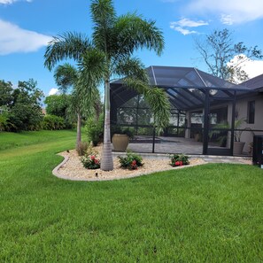 A covered outdoor patio area with a glass roof, surrounded by a well-maintained lawn and various plants.
