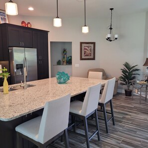 A modern kitchen with a granite countertop, stainless steel refrigerator, and pendant lighting.