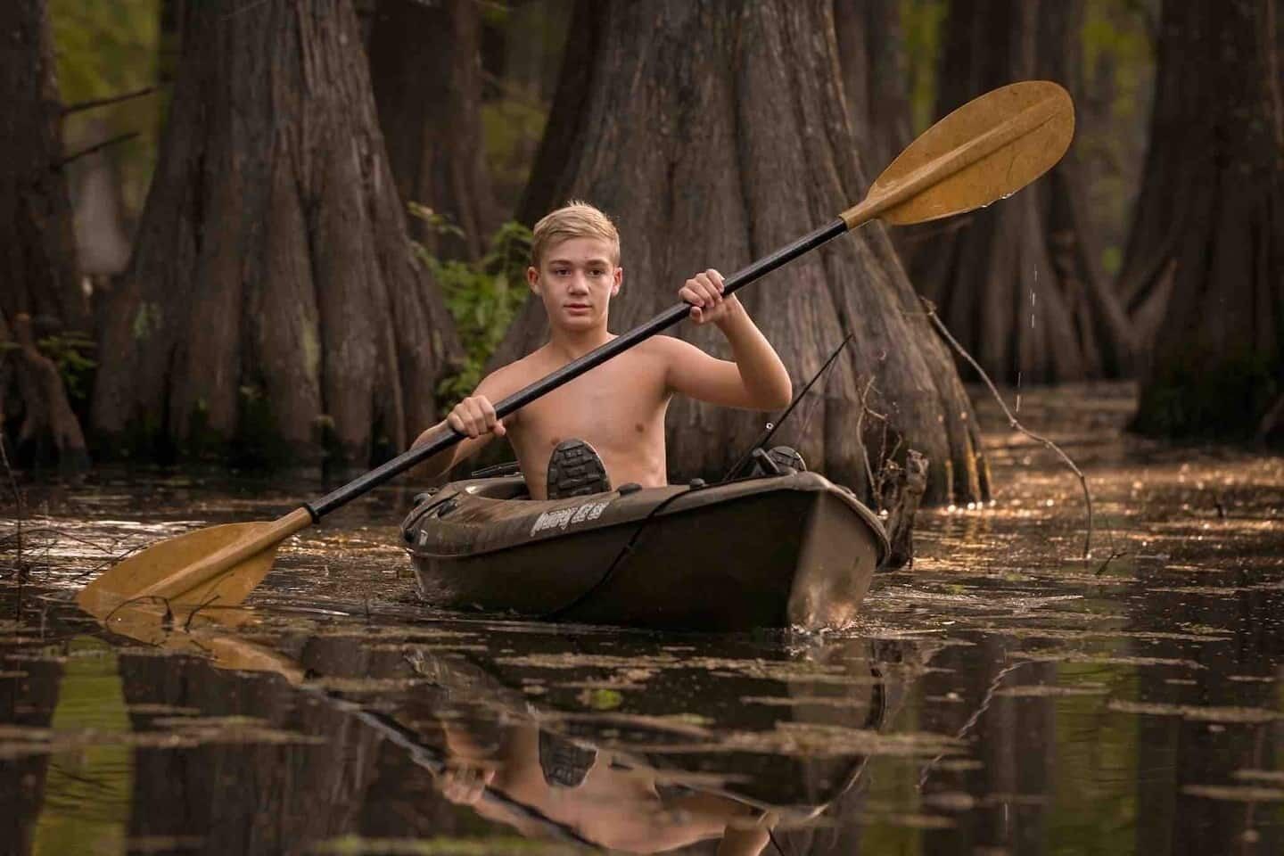 A person in a small boat with a paddle, surrounded by trees and water.