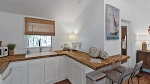 Una cocina con armarios blancos, una encimera de madera, una ventana con persiana de bambú, un fregadero, un microondas, una planta, un cuadro de un velero y un cubo de basura.