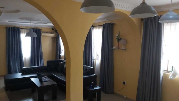 Sala de estar com sofá, mesa de centro e televisão. Cortinas nas janelas e um abajur pendurado no teto.