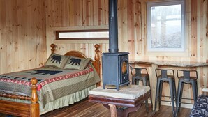 Una habitación tipo cabaña de madera con cama, estufa de leña, ventana y una pequeña mesa con taburetes.