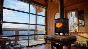 Interior acogedor de una cabaña con un amplio ventanal de madera, una chimenea de piedra y una terraza de madera con vistas a las montañas.