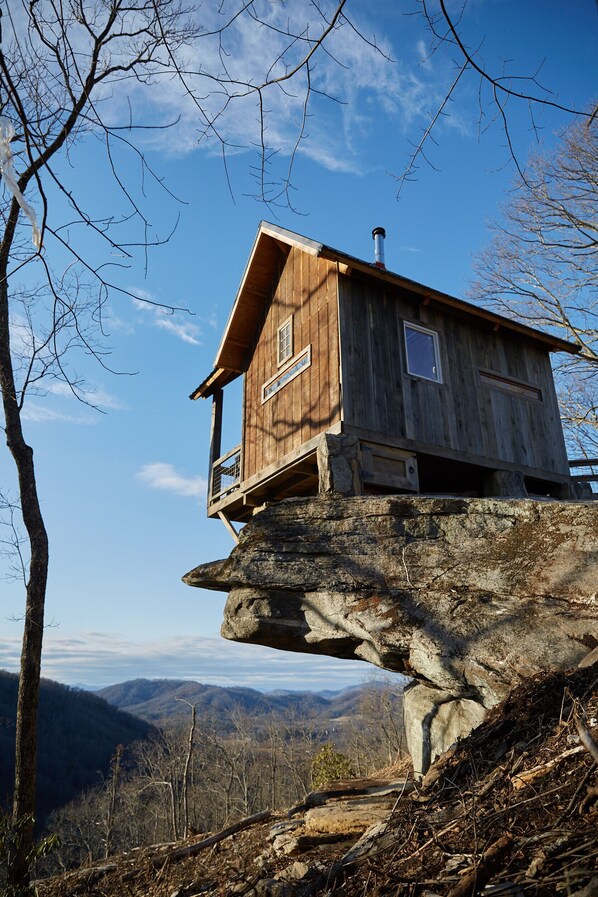 Una cabaña de madera sobre un saliente rocoso, con balcón y vista a un paisaje montañoso.