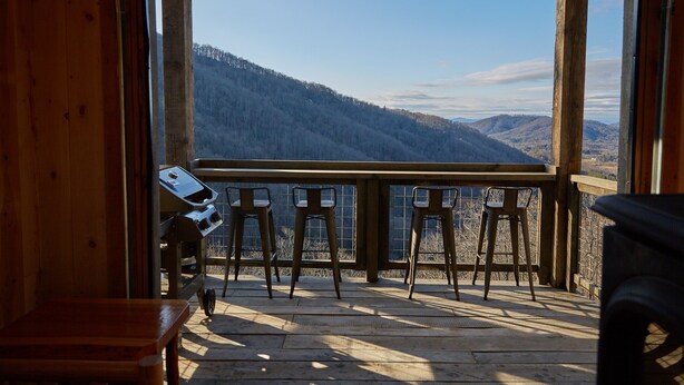 Una terraza de madera con una parrilla y taburetes de bar, con vistas a un paisaje montañoso.