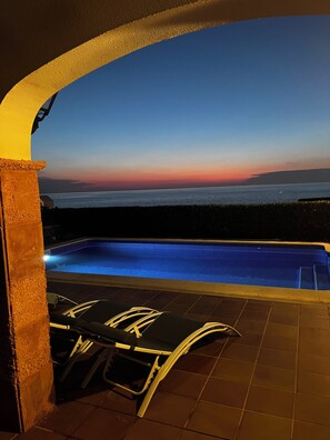 Una piscina al aire libre con vistas al atardecer, un área de estar cubierta y un patio empinado.