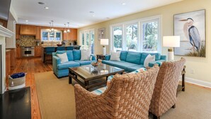 Un vaste salon avec un canapé bleu, une table basse et des fauteuils en osier. On aperçoit à l’arrière-plan une cuisine avec des meubles en bois et un coin repas comprenant une table et des chaises.