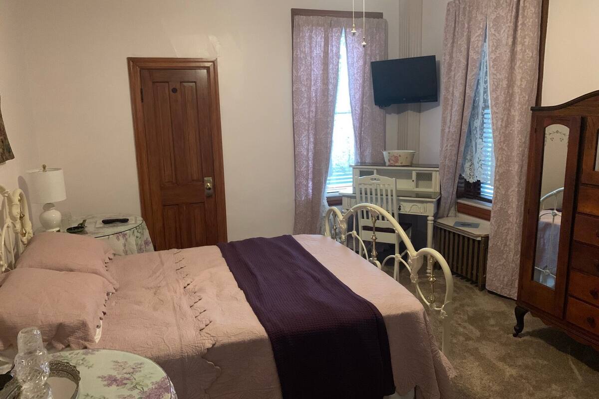 A bedroom with a bed, a dresser, a mirror, a television, and a window with curtains.