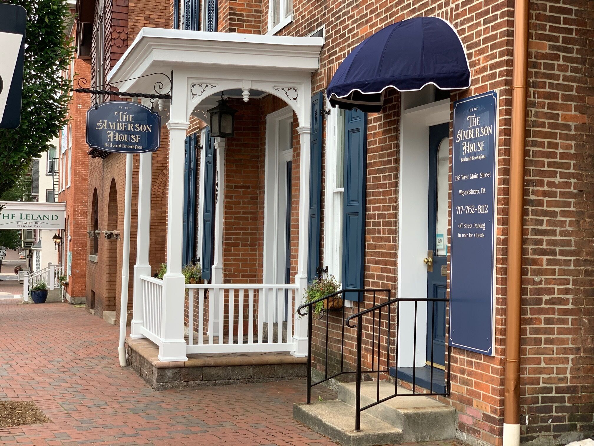 A brick building with a white porch and a blue awning. There are signs for 'The Amberson House' and 'The Leland'.