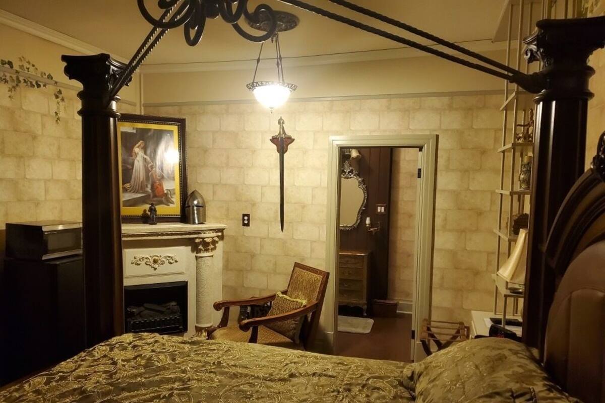 A bedroom with a four-poster bed, a fireplace, a chair, and a mirror.