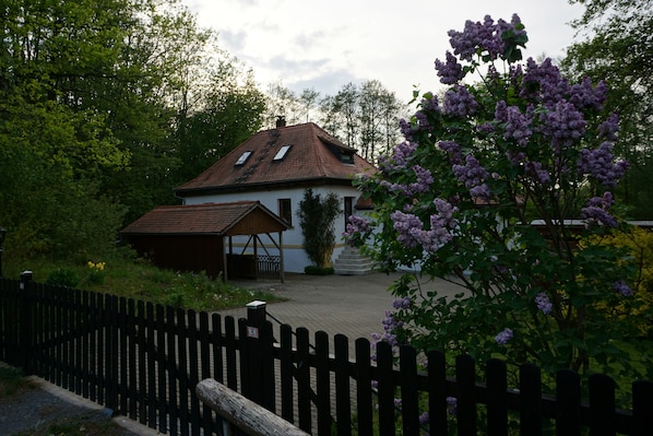 Una casa tradicional con techo de tejas, cerca de madera y un jardín con flores moradas.