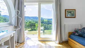 A modern bedroom with a wooden floor, a white desk with a chair, a bed with a blue bedspread, and a balcony with a view of green hills. - Beautiful private villa for 6 guests with A/C, WIFI, TV, patio and panoramic view (Casalborgone)