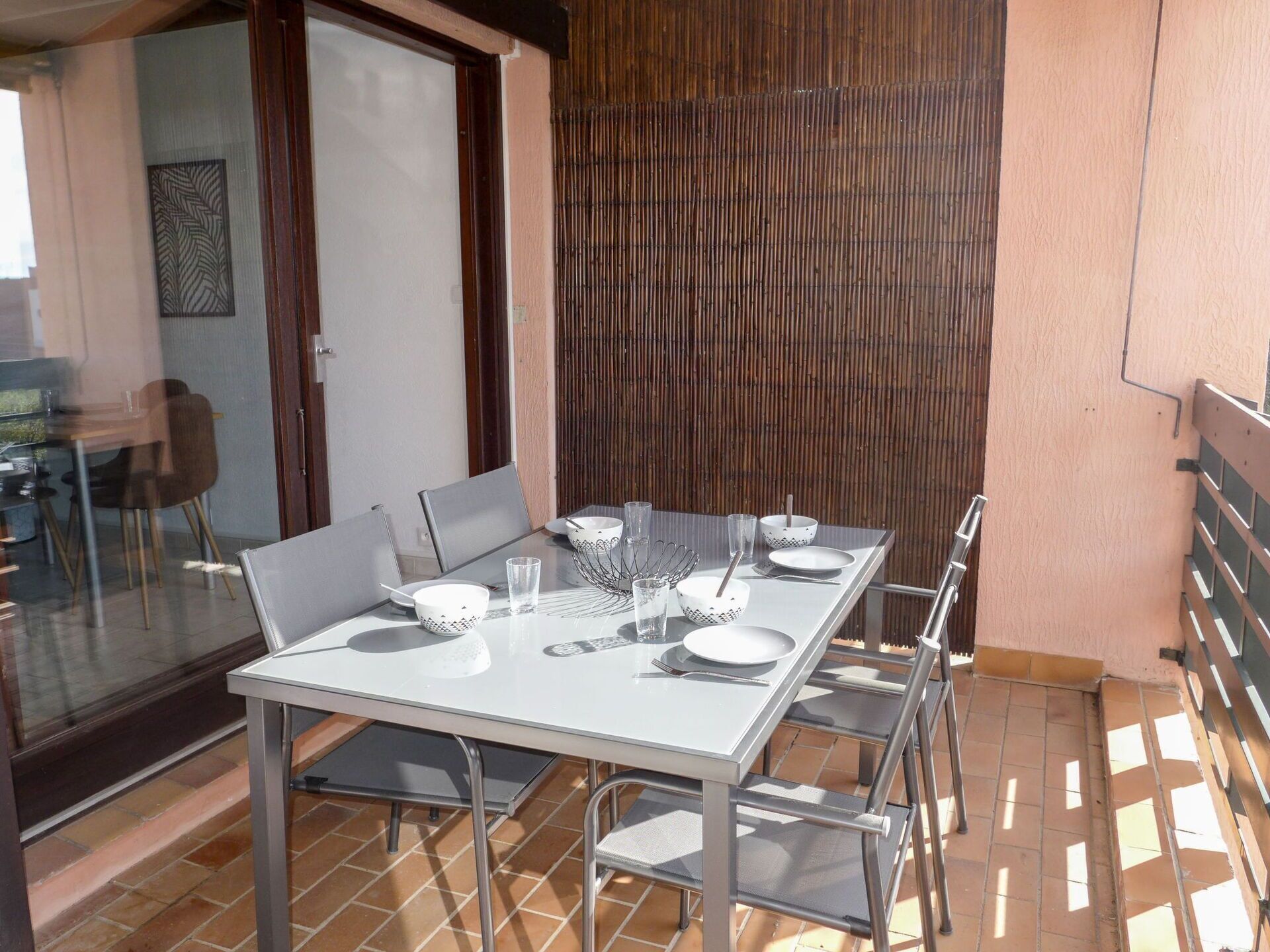 Un área de comedor al aire libre con mesa de vidrio y sillas de metal, un muro de separación de bambú y piso de baldosas.