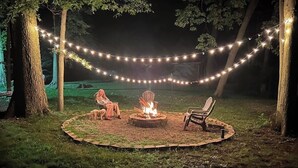 Un espace extérieur circulaire aménagé avec des sièges, doté d’un foyer, et entouré d’arbres et de guirlandes lumineuses.
