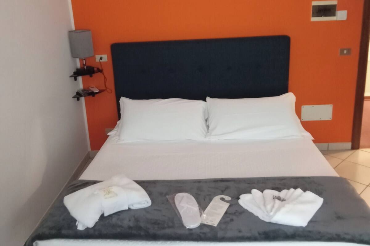 A hotel room with a bed, white pillows, a grey blanket, and a blue headboard. There is a lamp on the nightstand and a towel on the bed.