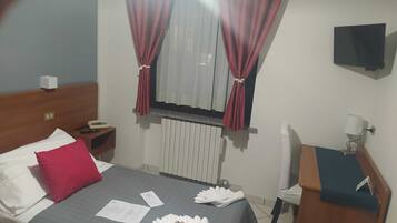 Ein Hotelzimmer mit Bett, Nachttisch, Schreibtisch, Stuhl, Fernseher und einem Fenster mit Vorhängen.
