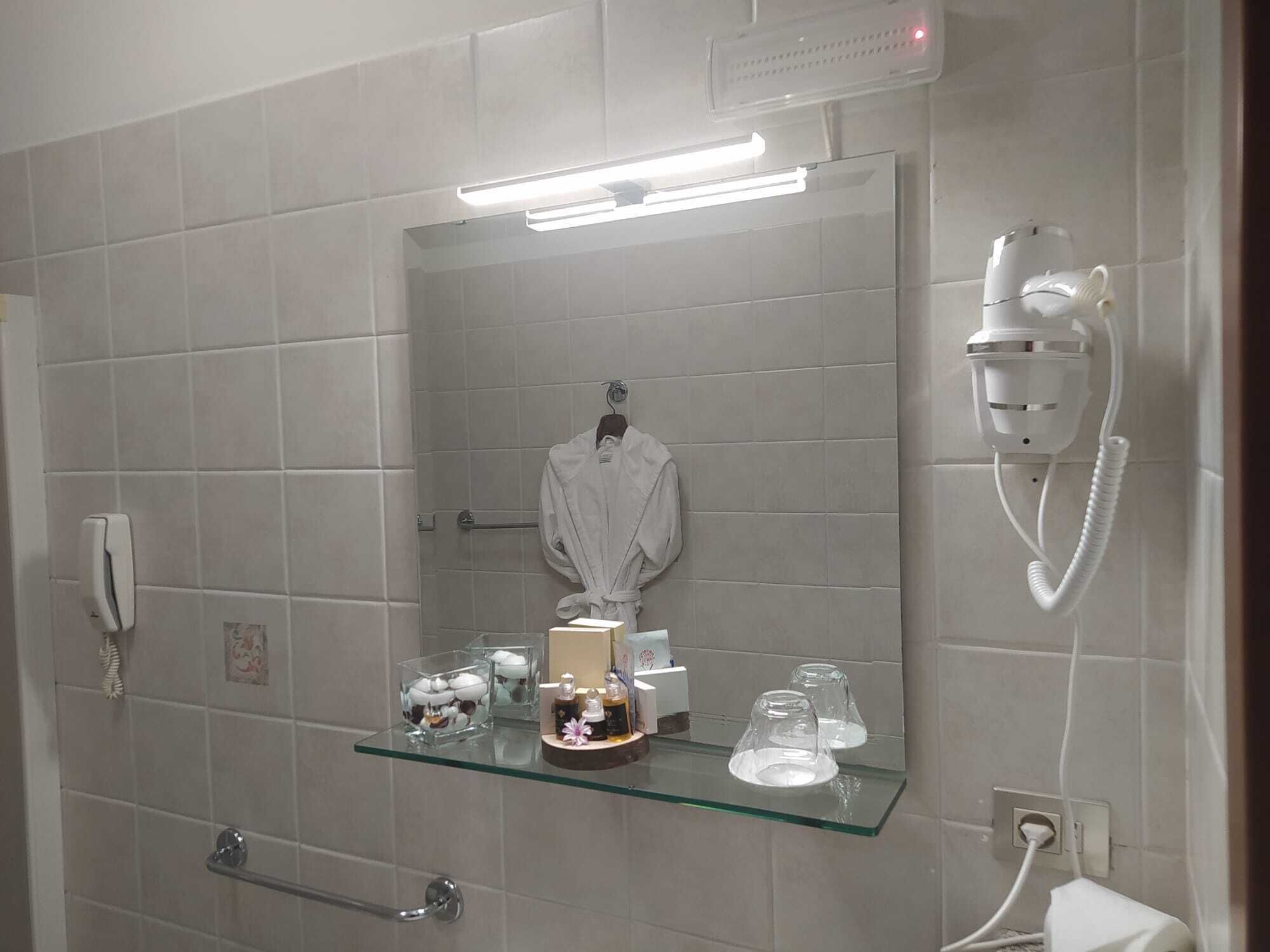 A bathroom with a glass shelf holding toiletries, a hairdryer mounted on the wall, and a towel rack with a robe.