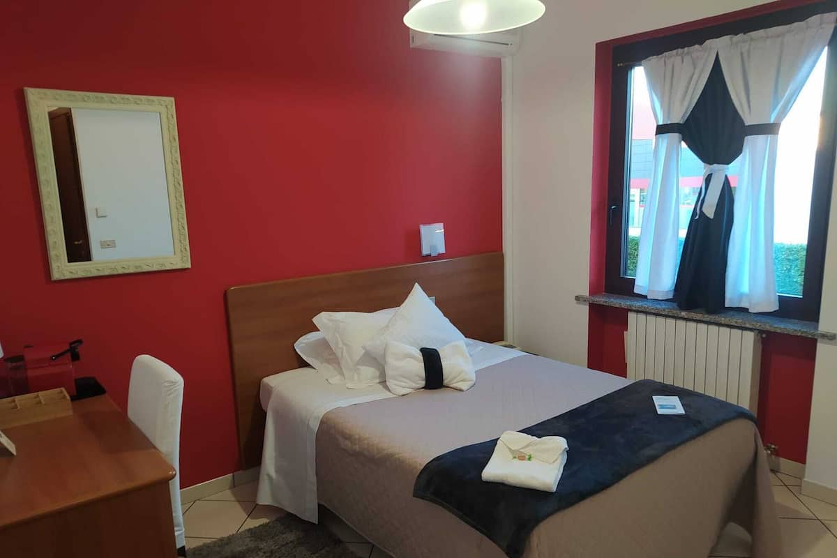 A hotel room with a bed, a desk, a chair, a window with curtains, and a radiator.