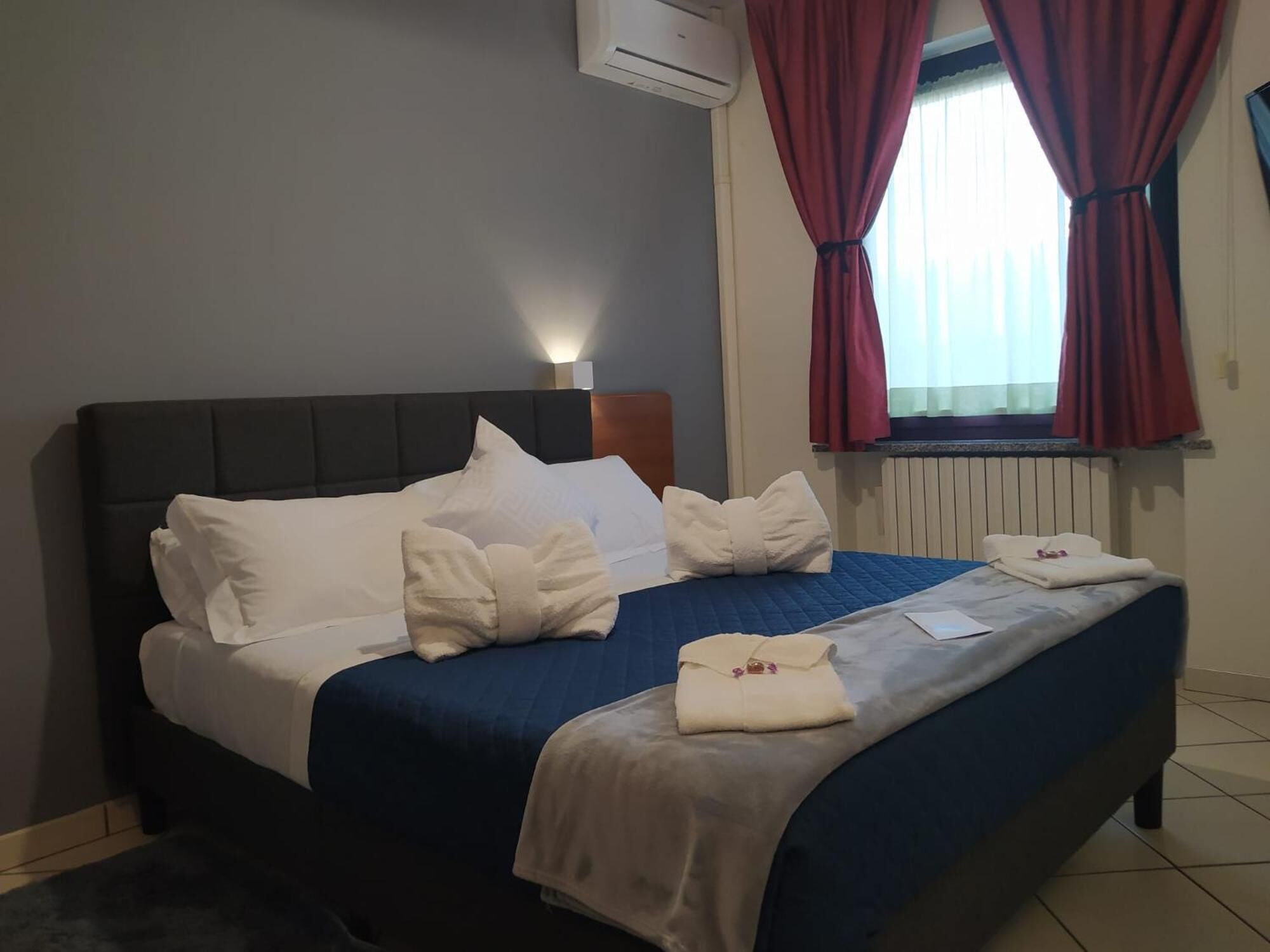 Un letto rifatto con lenzuola bianche e copripiumino blu, un condizionatore d'aria a parete e una finestra con tende rosse.