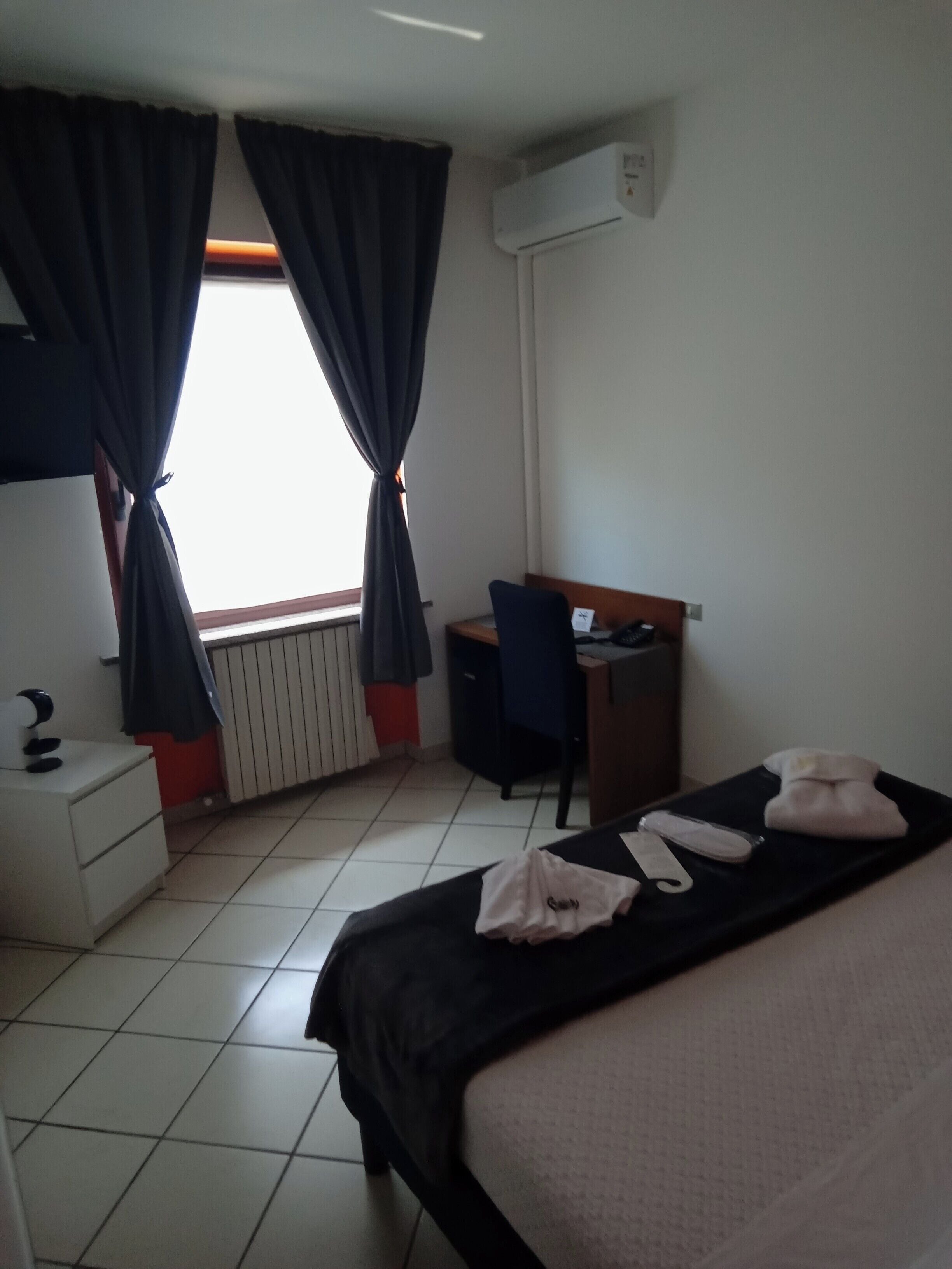 A hotel room with a bed, a desk, and a chair. There is a window with curtains, an air conditioning unit, and a radiator.