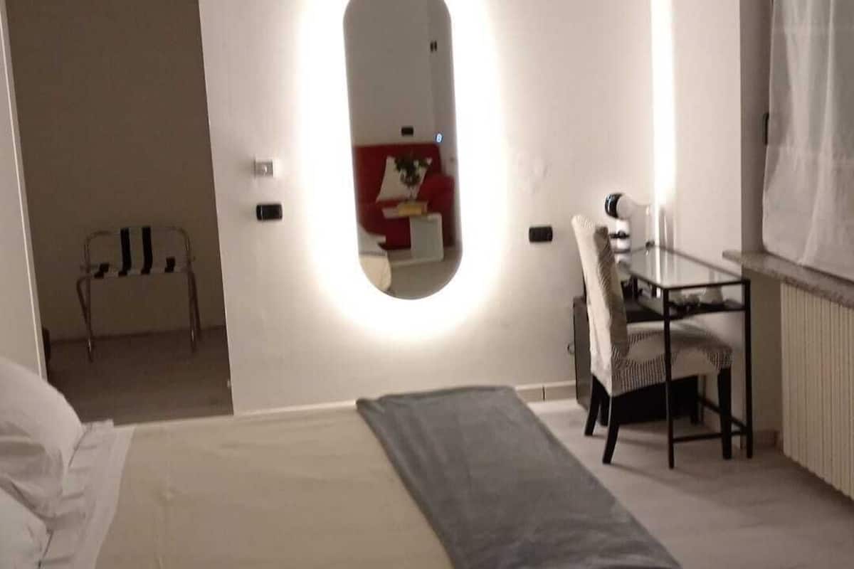 A modern bedroom with a bed, a desk, and a chair. There is a mirror on the wall and a small window.