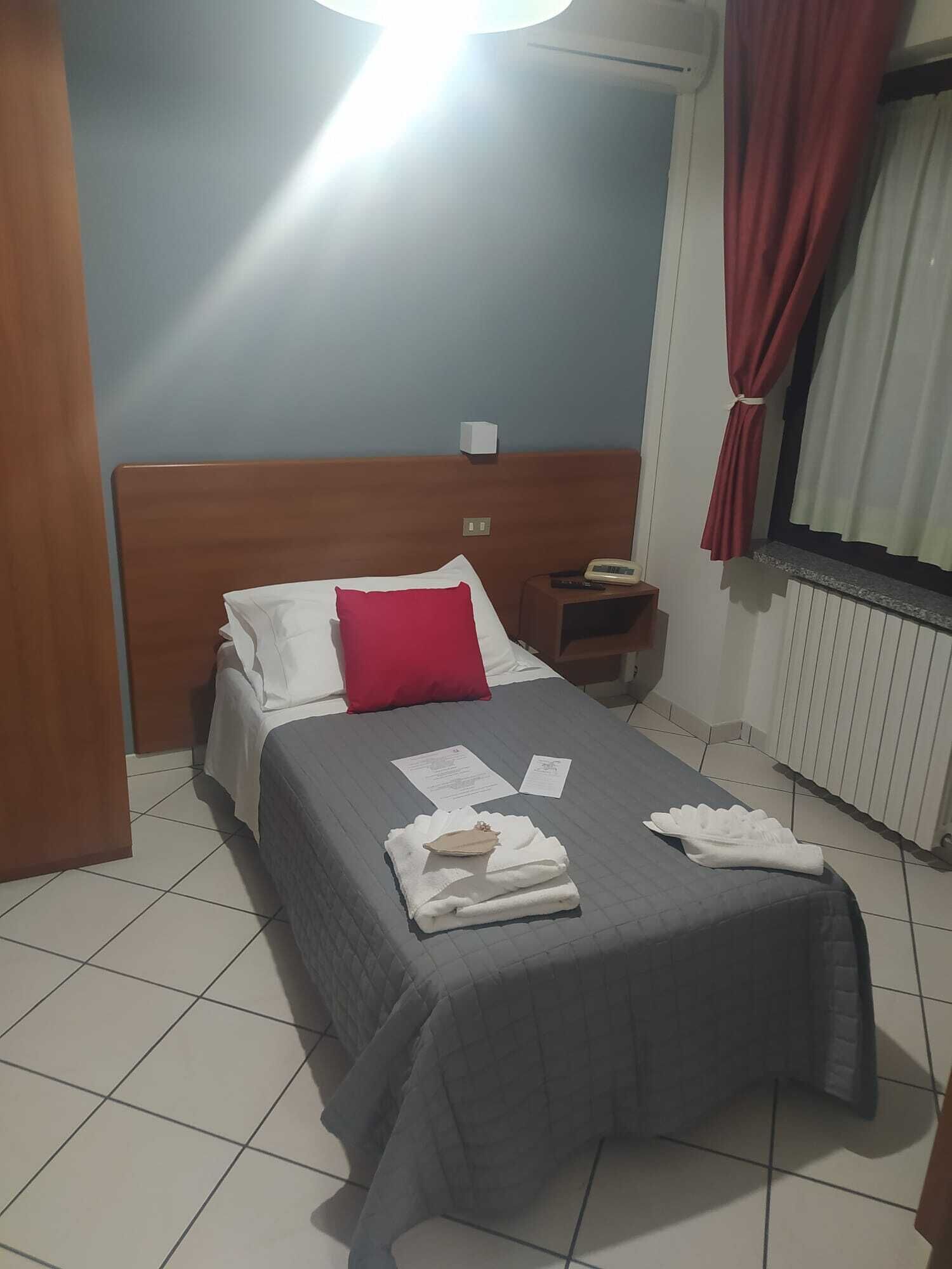 A hotel room with a bed, a nightstand, a red curtain, and a window with white curtains.