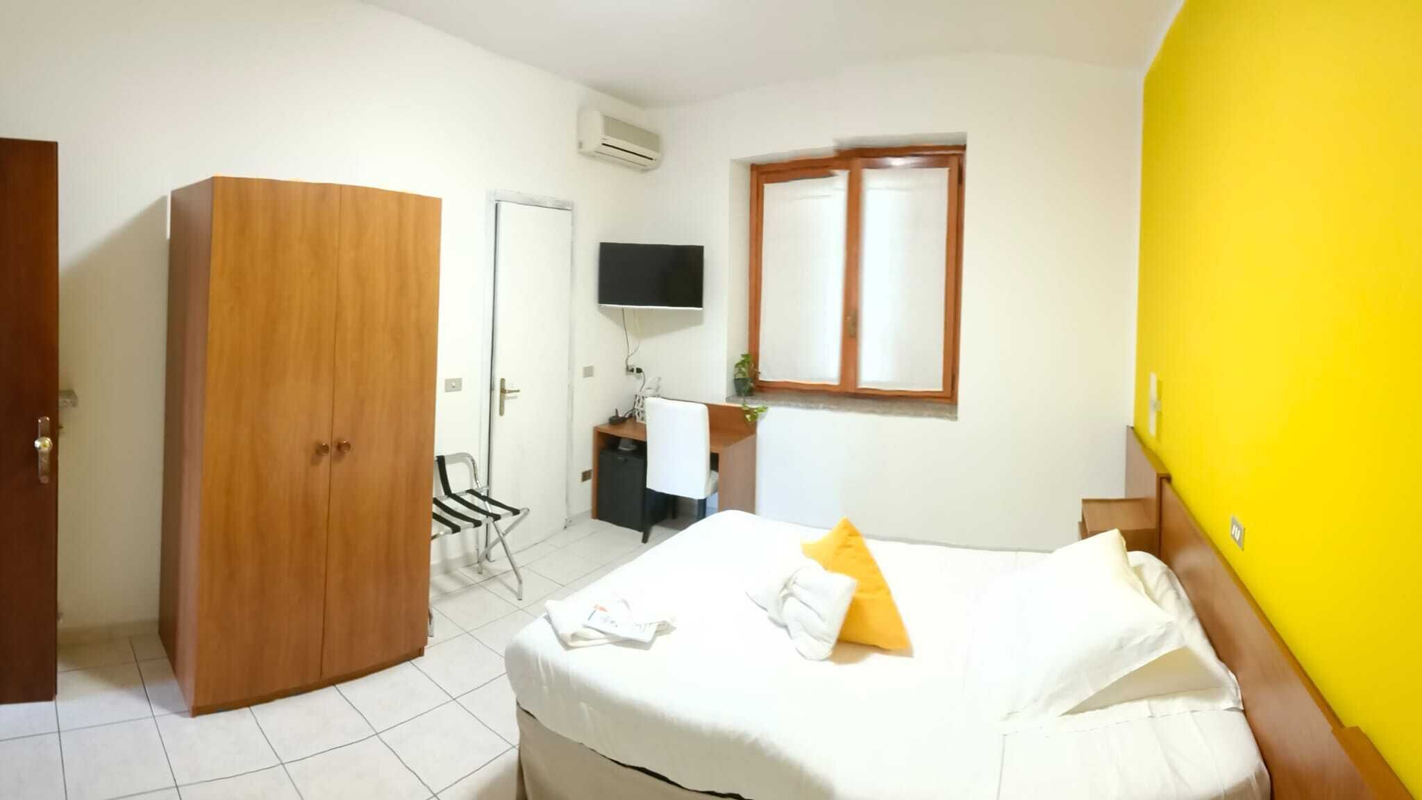 A hotel room with a bed, a wooden wardrobe, a desk with a chair, a television, and a window with a mirror.