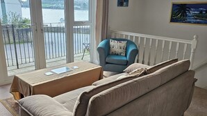A living room with a beige sofa, a blue armchair, and a view of a body of water through large windows. - Riverside retreat by Brunel’s Bridge, balcony view, off‑road parking, EV charger (Plymouth)