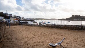 A sandy beach with a metal swing set and a metal chair. Boats are docked in the water behind a stone wall. - Riverside retreat by Brunel’s Bridge, balcony view, off‑road parking, EV charger (Plymouth)