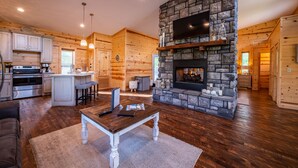 A spacious log cabin interior with a stone fireplace, wooden flooring, and a kitchen area with modern appliances. - Luxury Honeymoon cabin, jetted tub, tile shower, hot tub, fire pit (Broken Bow)