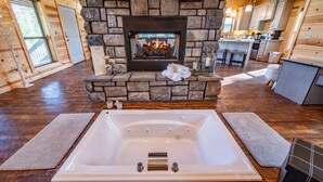 A cozy cabin interior with a stone fireplace, a large bathtub, and a kitchen area. - Luxury Honeymoon cabin, jetted tub, tile shower, hot tub, fire pit (Broken Bow)