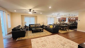 A spacious living room with a dark-colored sofa set, a coffee table, and a large floral rug. There is a dining area with a table and chairs, and a kitchen area with wooden cabinets and a microwave.