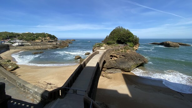A coastal view with a bridge leading to a beach, rocky outcrops, and a clear sky.