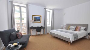 A hotel room with a bed, a sofa, a desk, and a chair. There is a window with curtains, a framed picture on the wall, and a small table with two cups and a bowl.