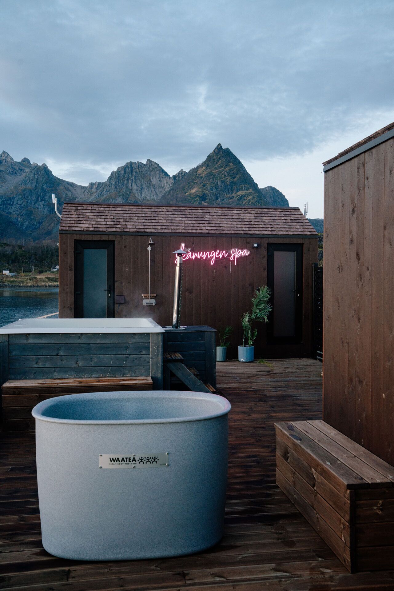 A spa with a wooden exterior, a neon sign, a hot tub, and a mountain backdrop.