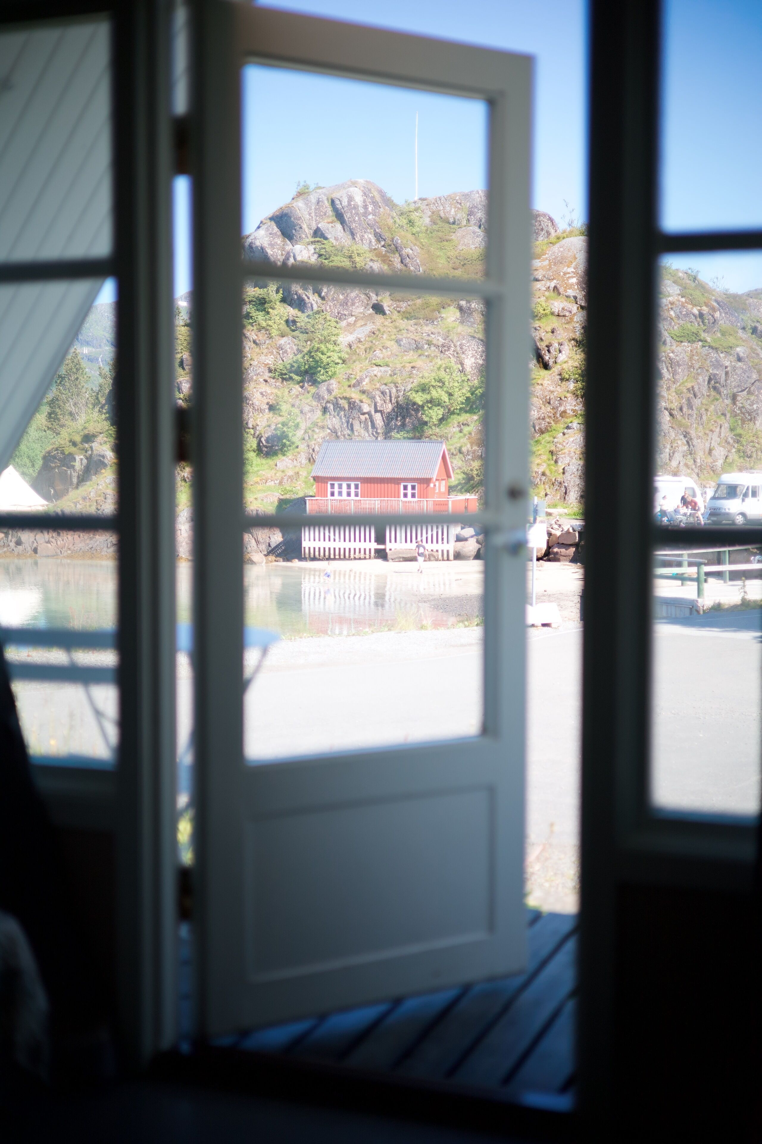 A view through a window of a building with a wooden deck, a small red building with a white fence, and a rocky hillside in the background.