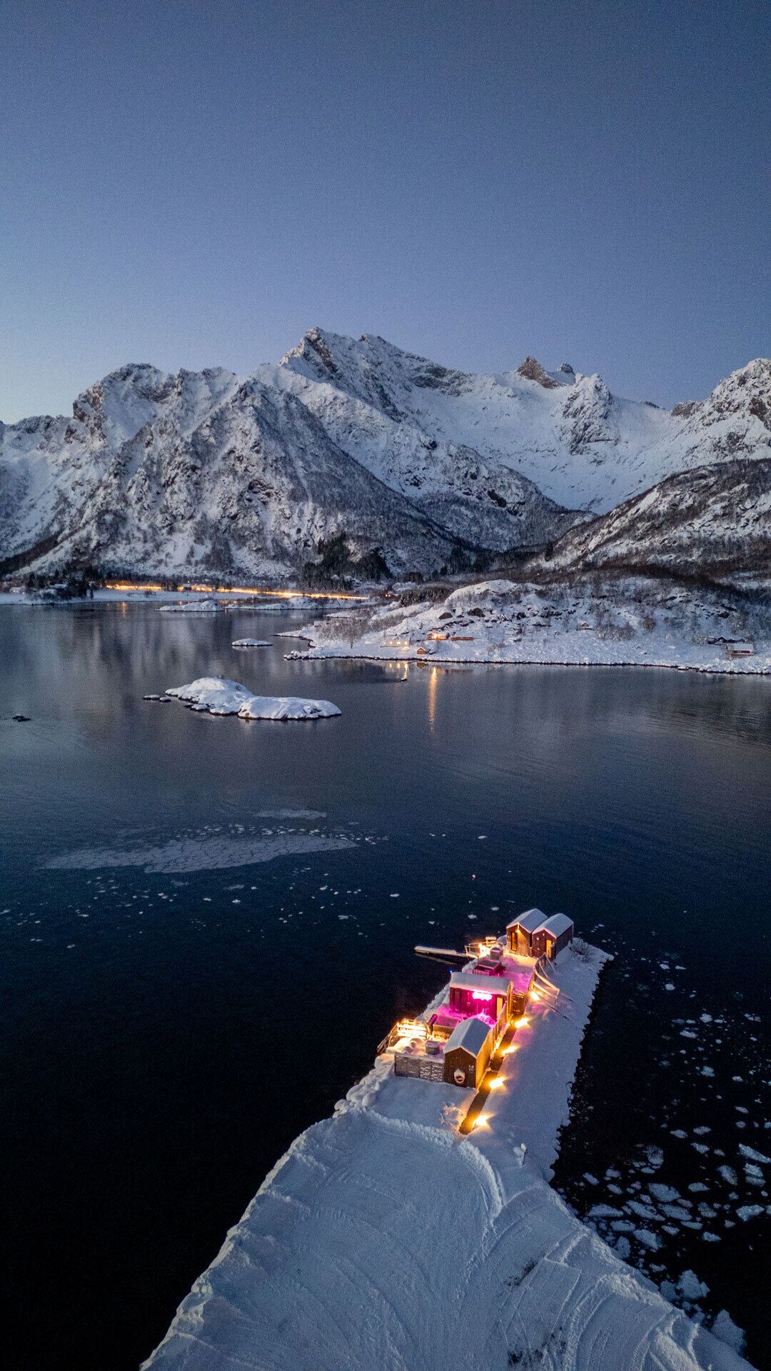 Paisaje nevado con un cuerpo de agua, icebergs y una cadena montañosa distante.