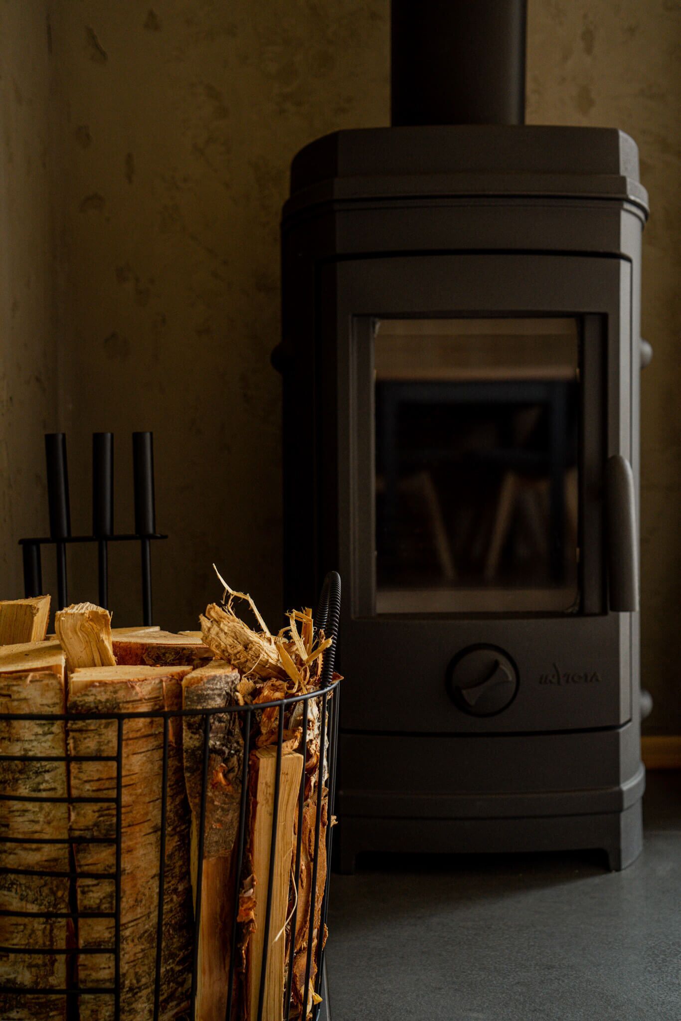 A wood-burning stove with a glass door, a metal rack with firewood, and a black stand with three vertical supports.