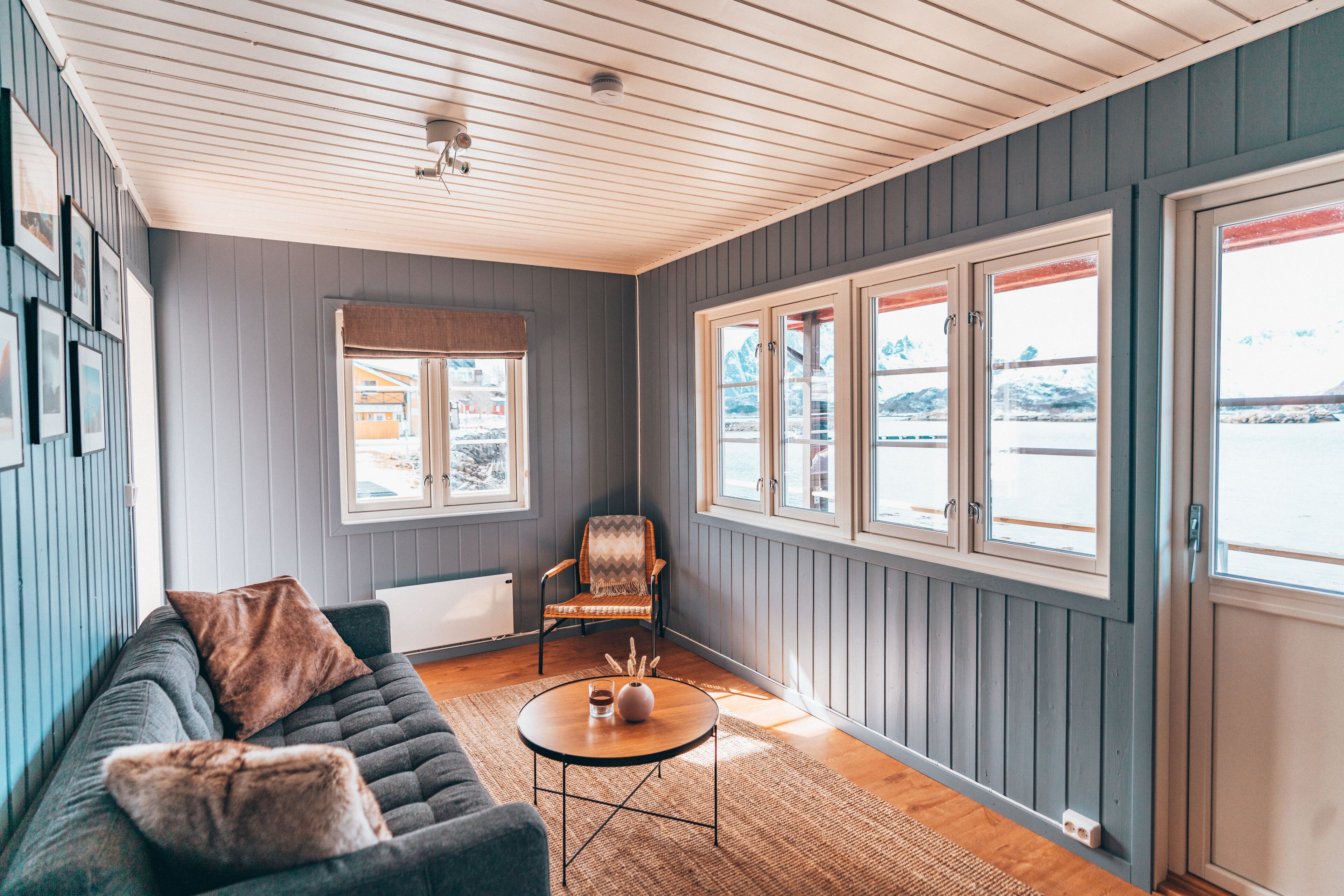 Un cuarto acogedor con un sofá gris, una mesa redonda para café y una silla. Hay ventanas amplias con marcos blancos y vistas al exterior.