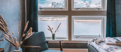 A room with a wooden floor, a blue chair, a bed, and a window with a view of snow-covered mountains.