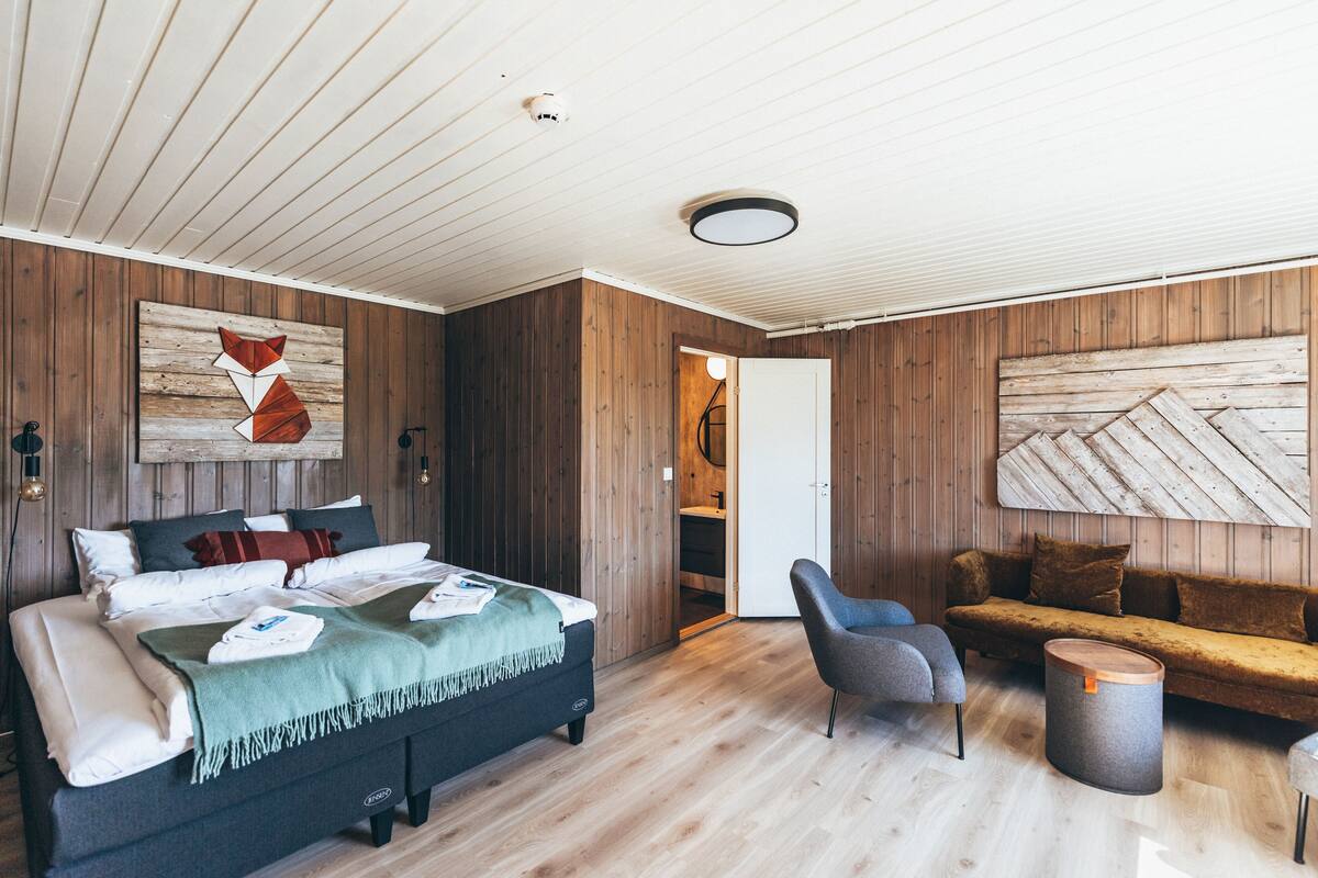 A modern bedroom with a wooden floor, a bed with multiple pillows, a grey armchair, a sofa, and a round wooden table.