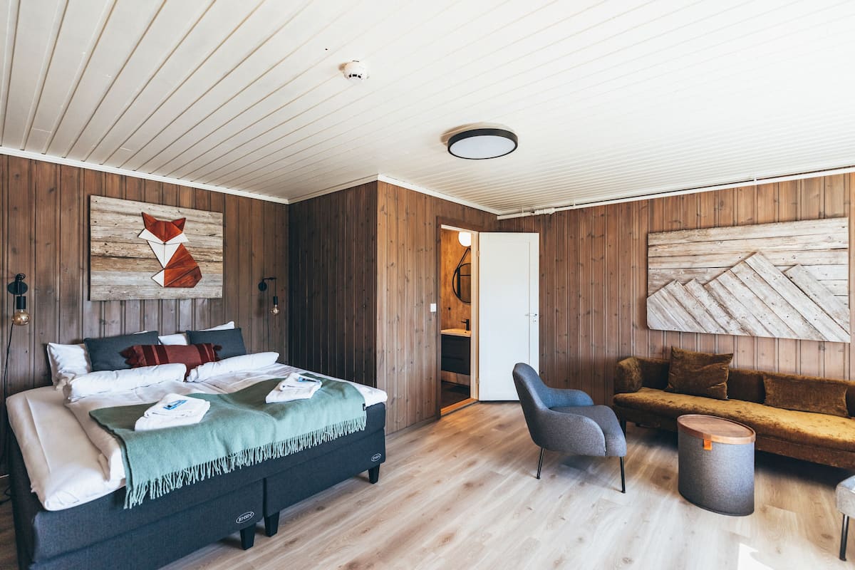 Un dormitorio moderno con piso de madera, una cama con muchos almohadones, un sillón gris, un sofá y una mesa redonda de madera.