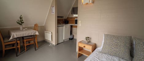 A compact room with a bed, a small table with a chair, a kitchenette, and a radiator.