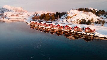 Una hilera de chalés de madera roja con tejados cubiertos de nieve, reflejándose en un lago tranquilo, rodeados de colinas y árboles nevados.